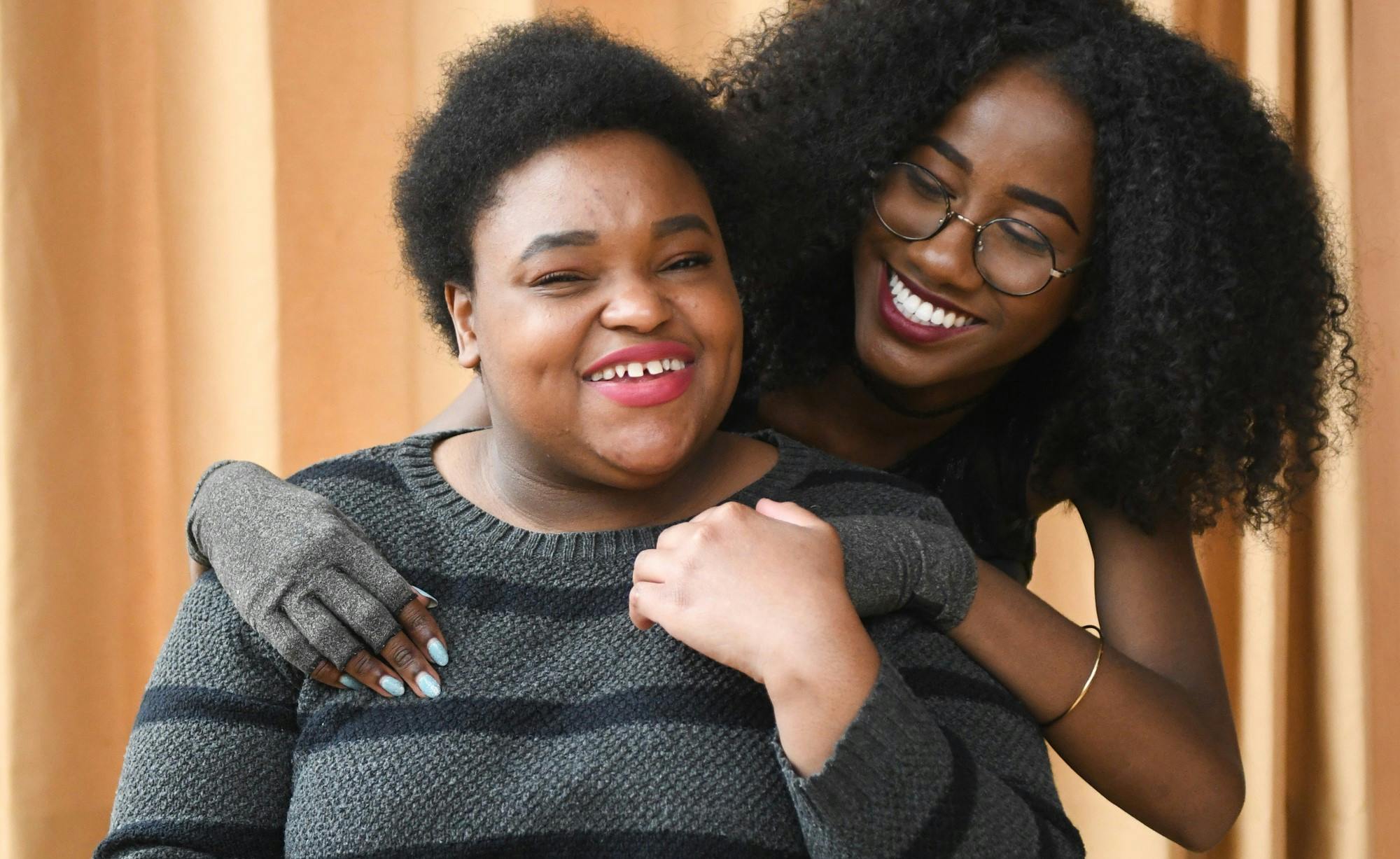 Close-up of a couple smiling and holding hands. The Black femme in the back wears compression gloves and looks lovingly at the non-binary Black person in the front, who sits in a power wheelchair. Photo attributed to Disabled and Here project.