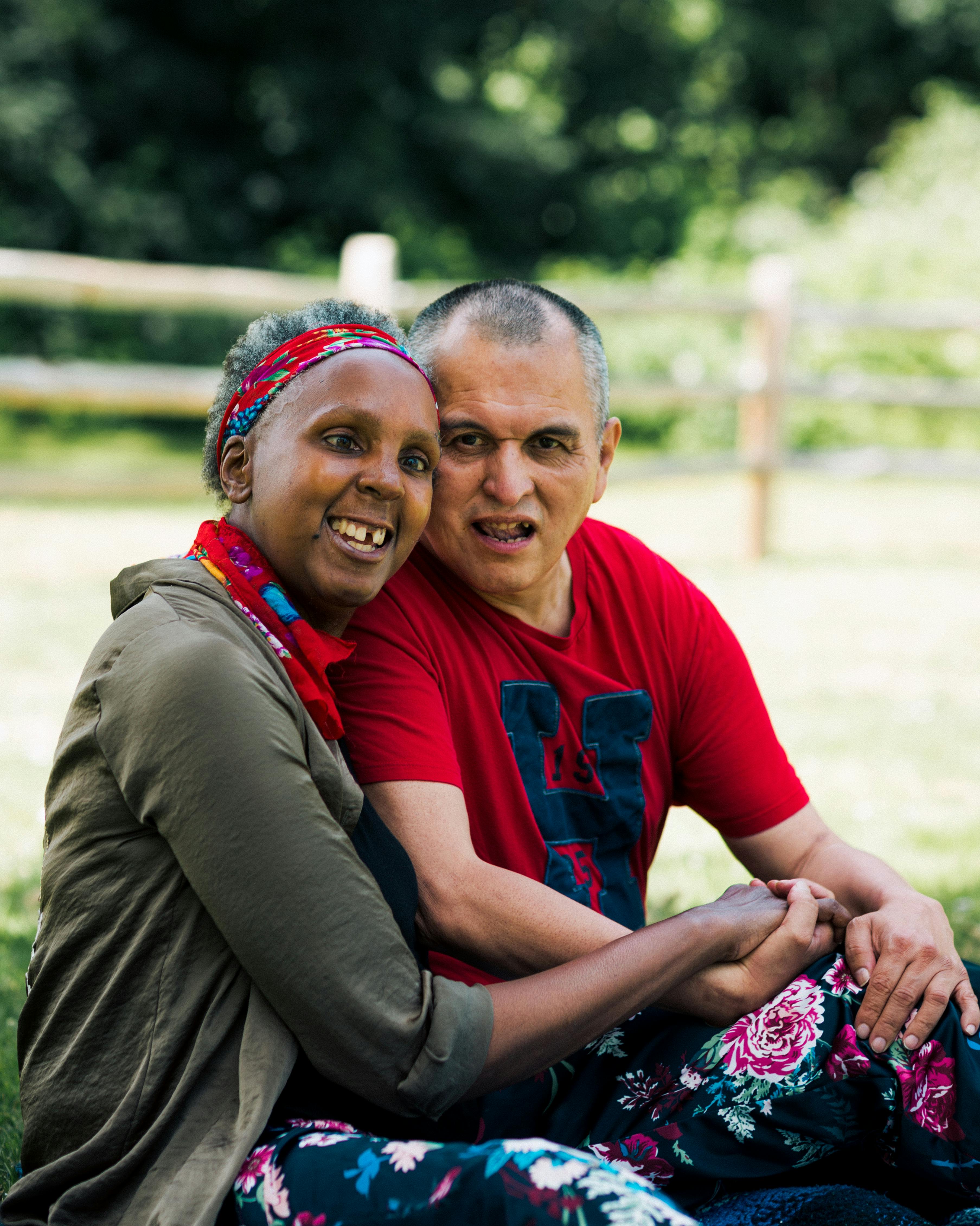 A disabled man and woman sitting on a blanket in the grass smiling,  holding hands and leaning their heads to touch together. The Black woman wears a read patterend headband,  dark green jacket and flower-printed skirt. The man wears a red shirt with the letter H printed in dark blue.