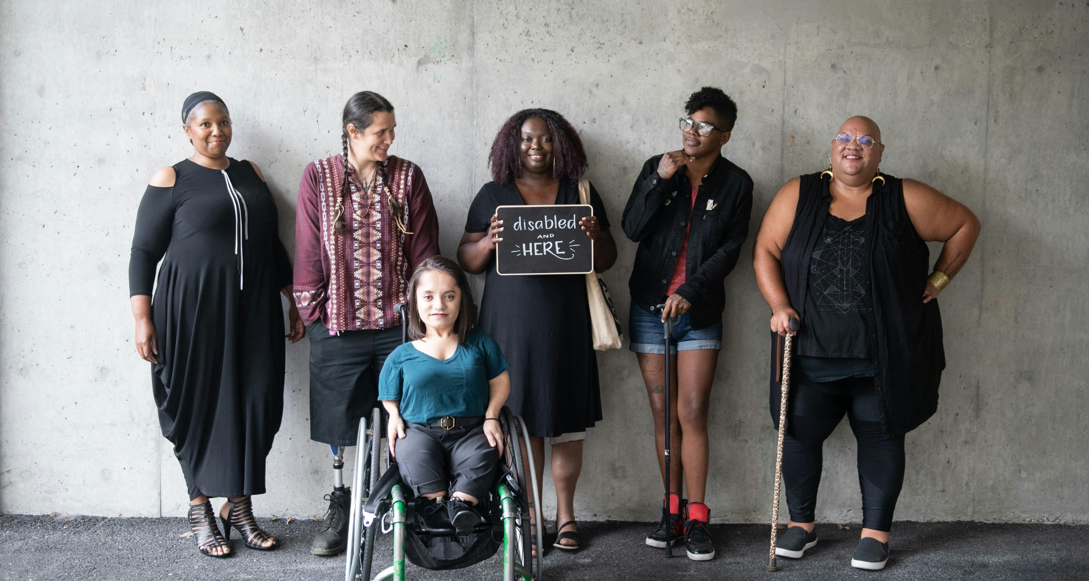 Six disabled people of color smile and pose in front of a concrete wall. Five people stand in the back, with the Black woman in the center holding up a chalkboard sign reading `disabled and here.` A South Asian person in a wheelchair sits in front. Photo attributed to Disabled and Here project.