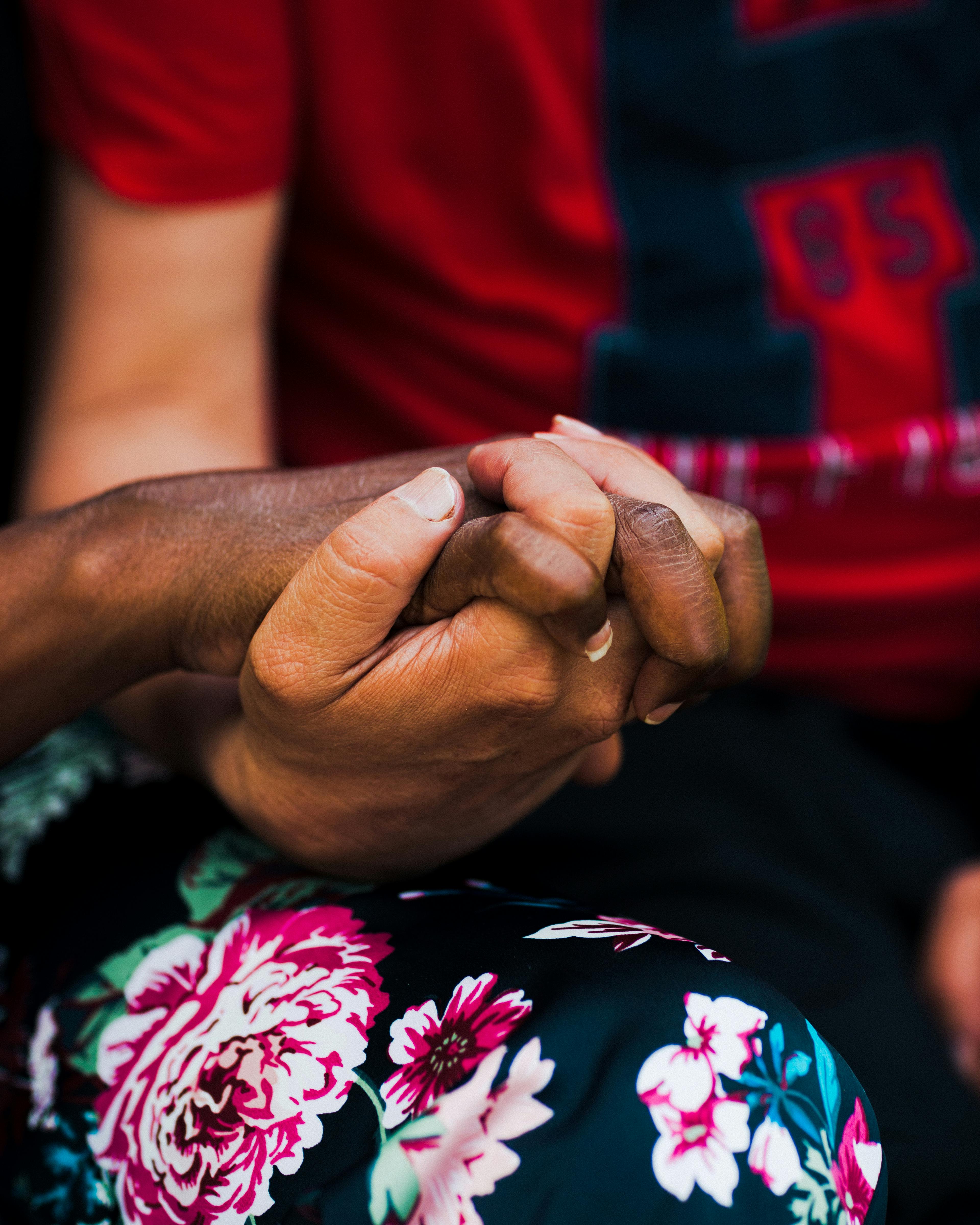 Close up of disabled couple holding hands with interlaced fingers. A Black woman wears a floral printed skirt,  and the man wears a red shirt with the letter H printed in dark blue.